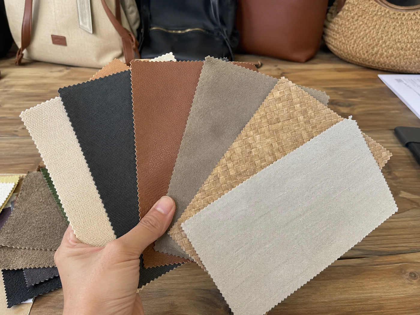 Smartphone-style buyer hand comparing canvas, nylon, PU, suede-like, raffia, and lining swatches on a sample table