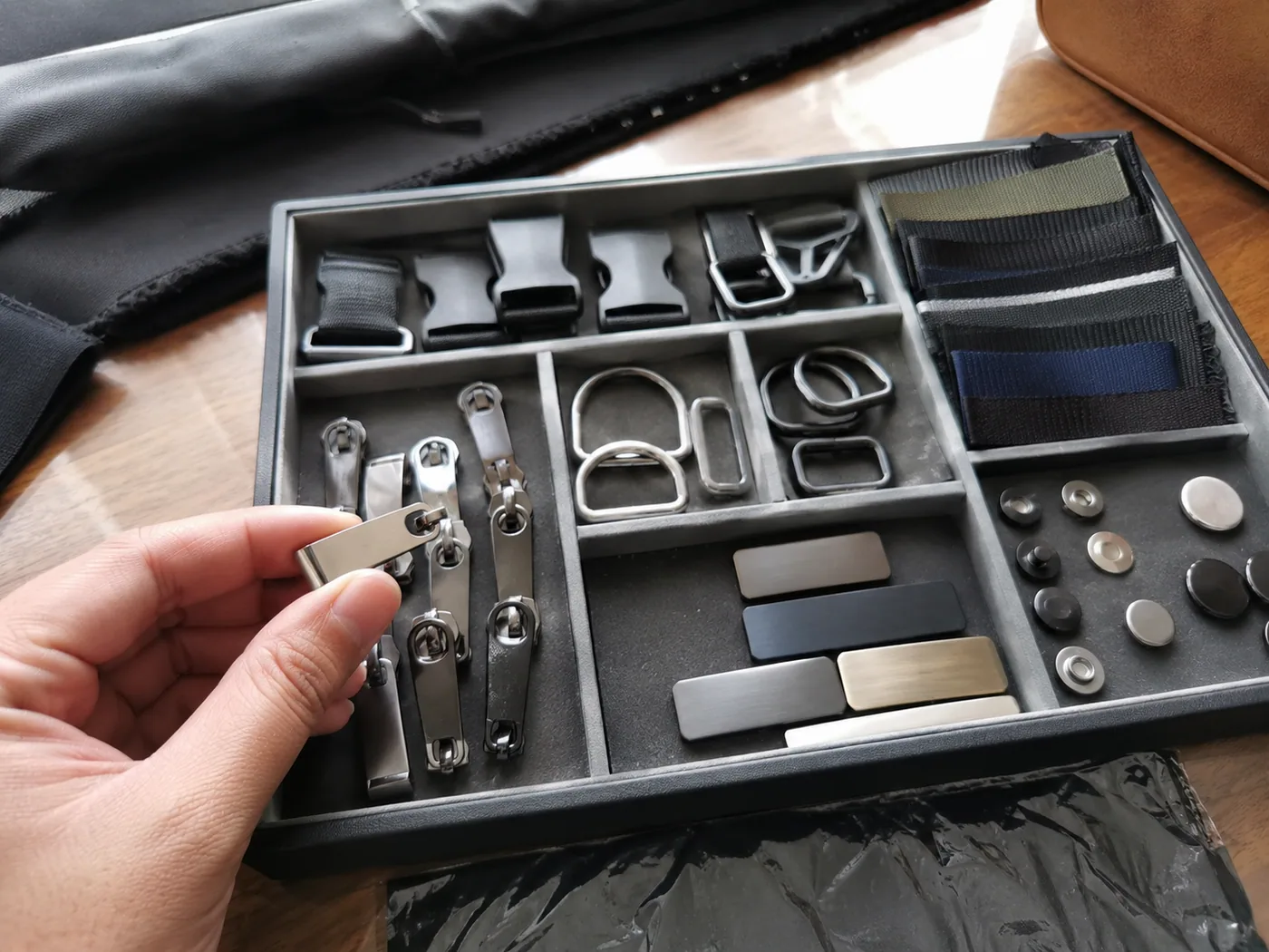 Smartphone-style close-up of zipper pullers, buckles, D-rings, webbing, snap buttons, and blank logo plates on a QC desk